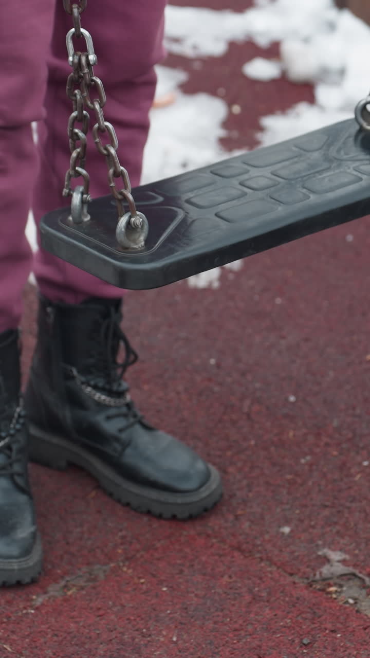 vista de la pierna de una persona arrodillada en el columpio con la rodilla derecha colocada antes de la izquierda, con botas negras en el suelo de textura roja con manchas de nieve