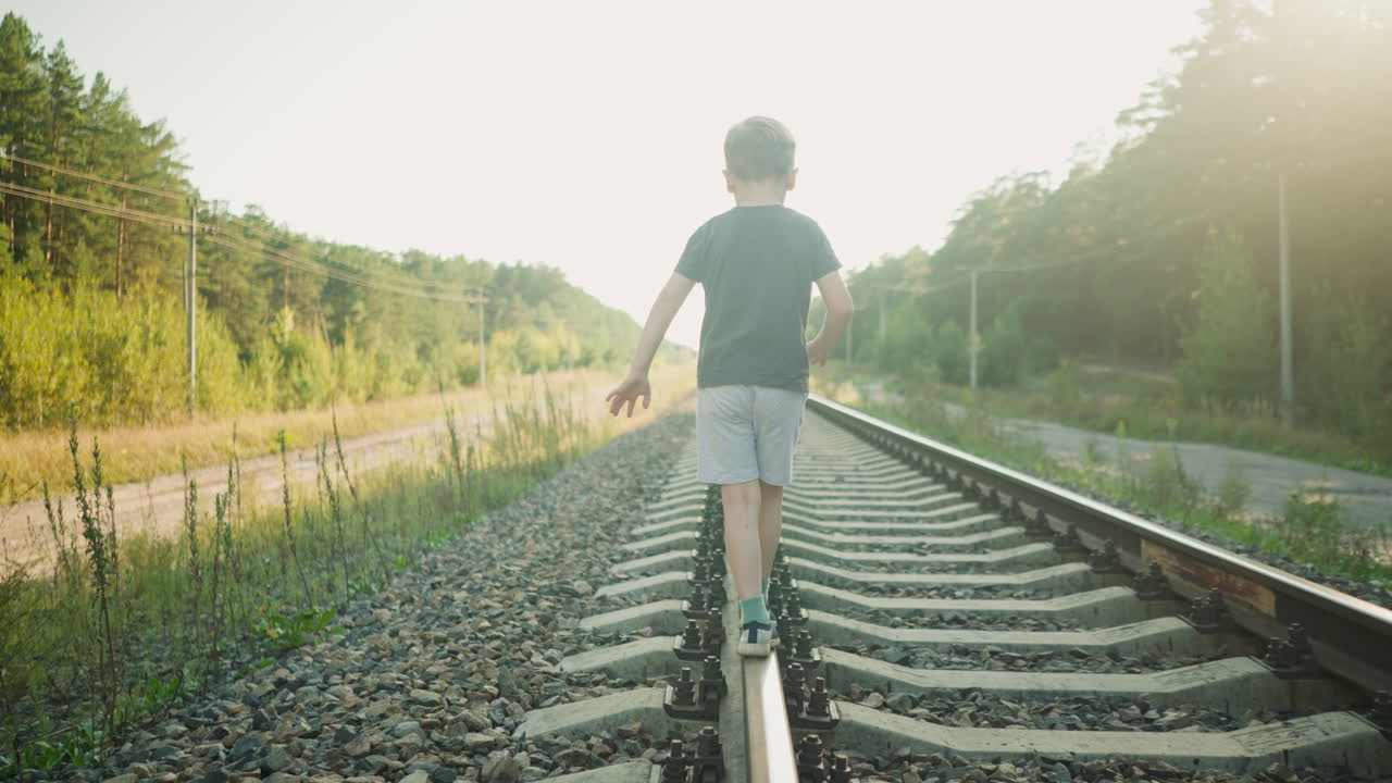 back view of kid walking carefully along rail track trying to balance with arms stretched out, surrounded by gravel, forest, and power lines under morning light creating soft glowing effect