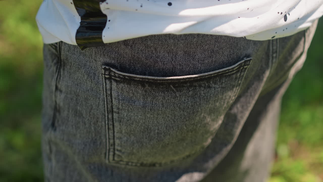 Primo piano all'aperto di un adolescente di colore con tasca in denim nera. La mano scivola nella tasca posteriore vicino allo smartwatch, jeans grigi testurizzati, cuciture e dettagli delle cuciture visibili, sfondo di erba illuminata dal sole, atteggiamento casual e sicuro di sé
