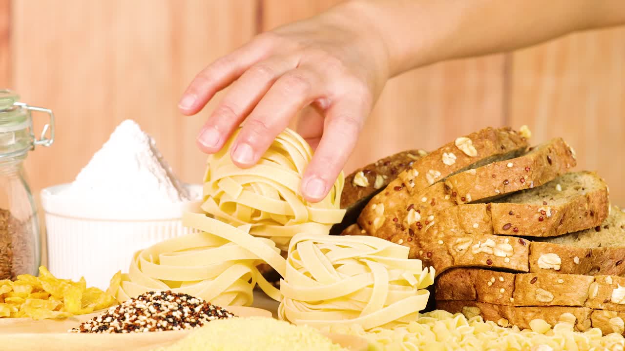 A hand selects tagliatelle from a display of bread and grains. Bright lighting, wooden background, and a warm, inviting atmosphere