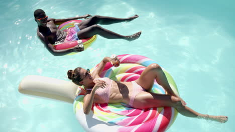 relaxing on pool floats, diverse couple enjoying drinks and sunshine in swimming pool