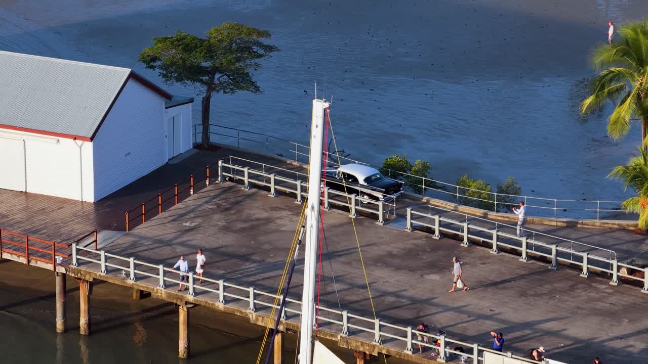 Drone footage captures people strolling along a tropical marina jetty in Port Douglas, Queensland, under clear skies and bright sunlight