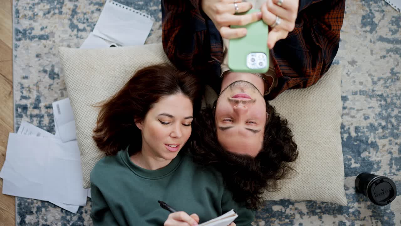 행복한 갈색 소녀가 노트북에 메모를 하고 그녀의 남자친구는 곱슬 머리카락으로 현대 아파트의 바닥에 베개에 누워있는 소셜 네트워크를 스크롤합니다.