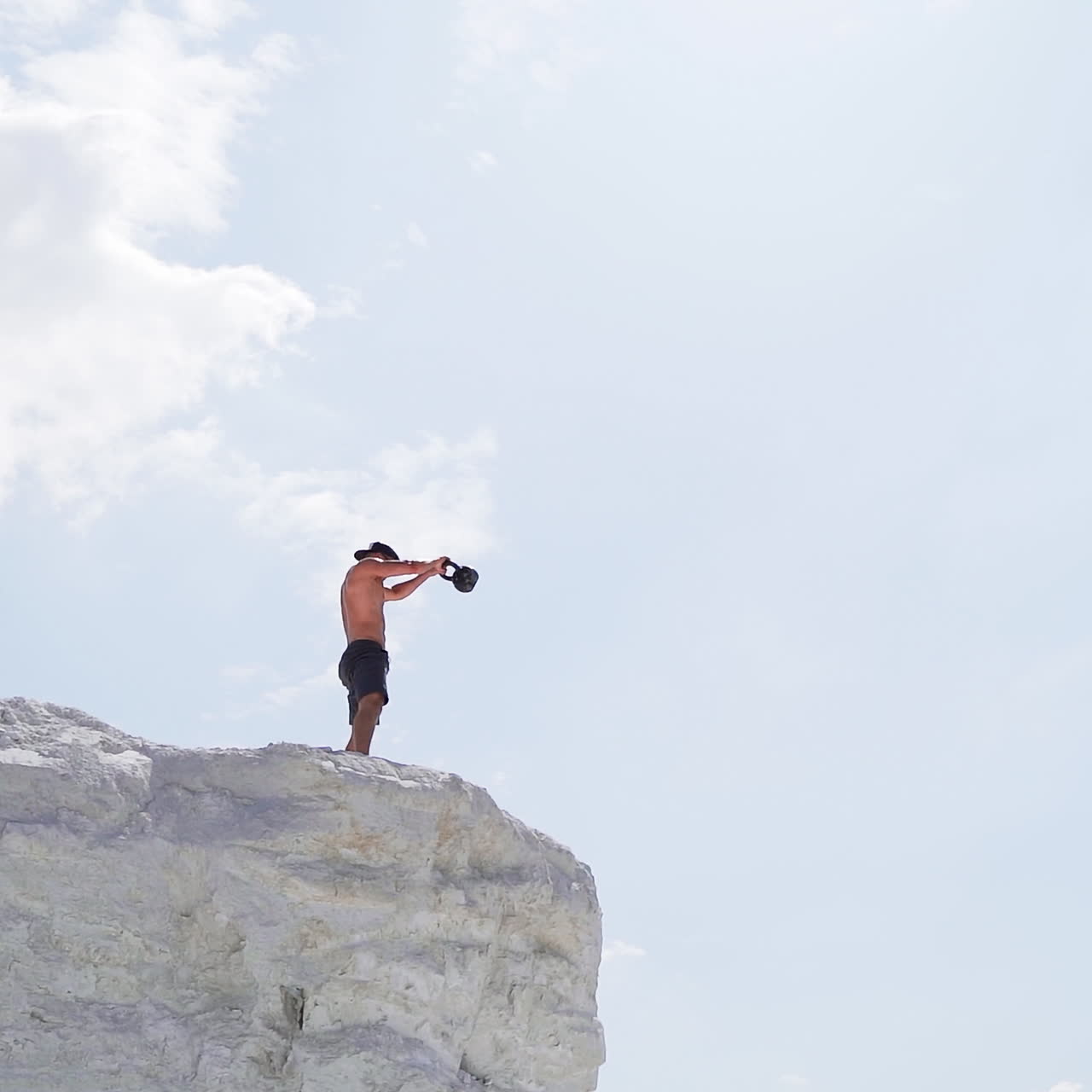View from below on a healthy man training with heavy weight on a hill. Bodybuilder lifting kettlebell on the natural sky background. Shirtless man during workout outdoors.
