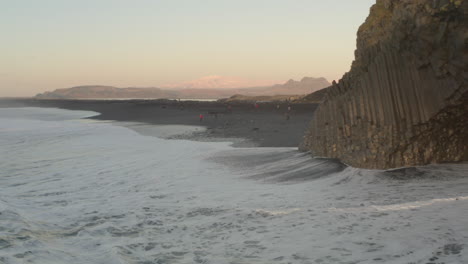 Toma-Aérea-Hacia-Los-Turistas-En-La-Playa-De-Arena-Negra-Columnes-Reynisfjara-Islandia