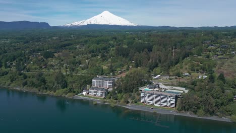 ascending drone shot over lake and modern waterfront buildings with snow-capped villarrica volcano in the background surrounded by lush forest
