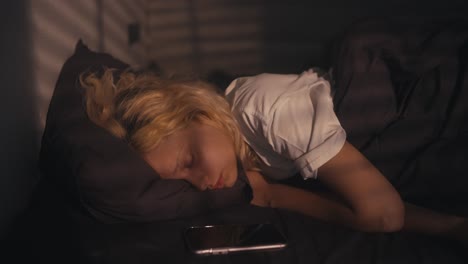 Tired-blonde-girl-in-a-white-T-shirt-sleeps-and-is-woken-up-by-an-alarm-clock-early-in-the-morning-in-a-gray-bed-at-home