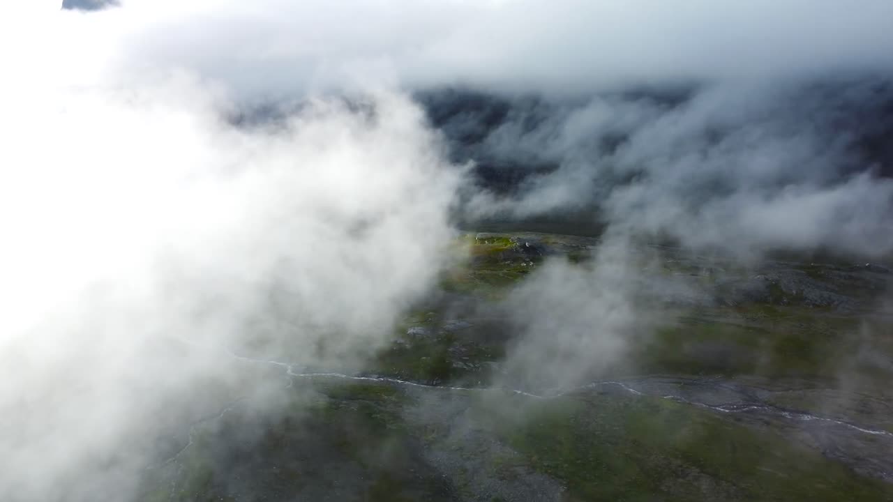 Aerial drone footage flying through thick white and fluffy clouds above Sweden green gorgeous landscapes that have a small narrow river flowing on it during day time.