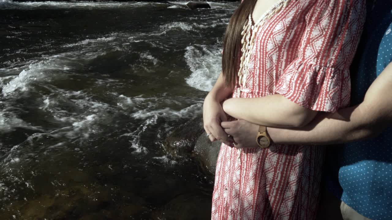 울창하고 푸른 삼림을 흐르는 물줄기 옆에 서 있는 아내를 뒤에서 껴안고 있는 남자