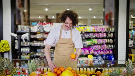 Confident-brunette-supermarket-worker-with-curly-hair-in-an-apron-lays-out-citrus-and-orange-fruits-on-a-shelf-in-a-supermarket