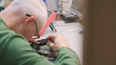 electronic equipment repair shop. the engineer technician solders the printed circuit board of an electronic device under a microscope.