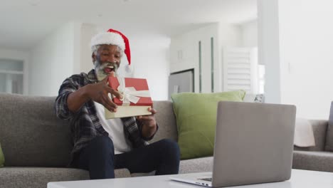 senior man wearing santa hat opening gift box while having video chat on his laptop