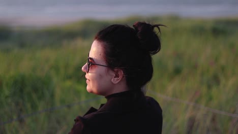 chica con gafas de sol viendo la puesta de sol junto al océano mientras gira la cabeza y se sienta en un banco