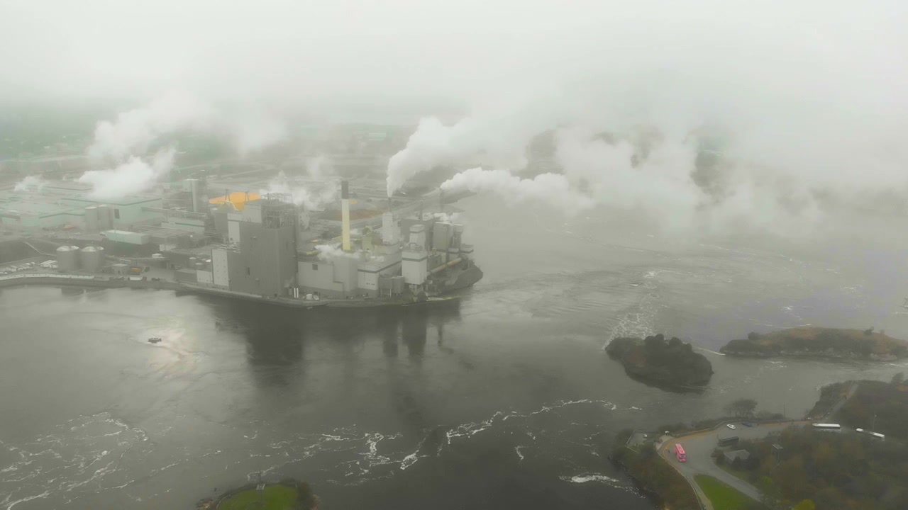 Aerial shot emerging from the thick clouds to reveal the Irving Pulp and Paper plant next to the reversing falls as it shoots toxic smoke into the air.