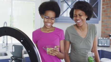 Happy-african-american-mother-and-daughter-preparing-healthy-drink,-making-video-using-smartphone