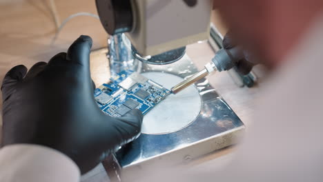 behind view of a technician's gloved hands meticulously soldering a blue circuit board under a microscope, the technician uses a soldering iron to work on the circuit