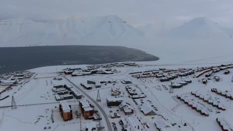 Vista-De-Drones-En-Svalbard-Volando-Sobre-La-Ciudad-De-Longyearbyen-Mostrando-Casas-En-Una-Zona-Nevada-Con-Un-Fiordo-Y-Una-Montaña-En-Noruega