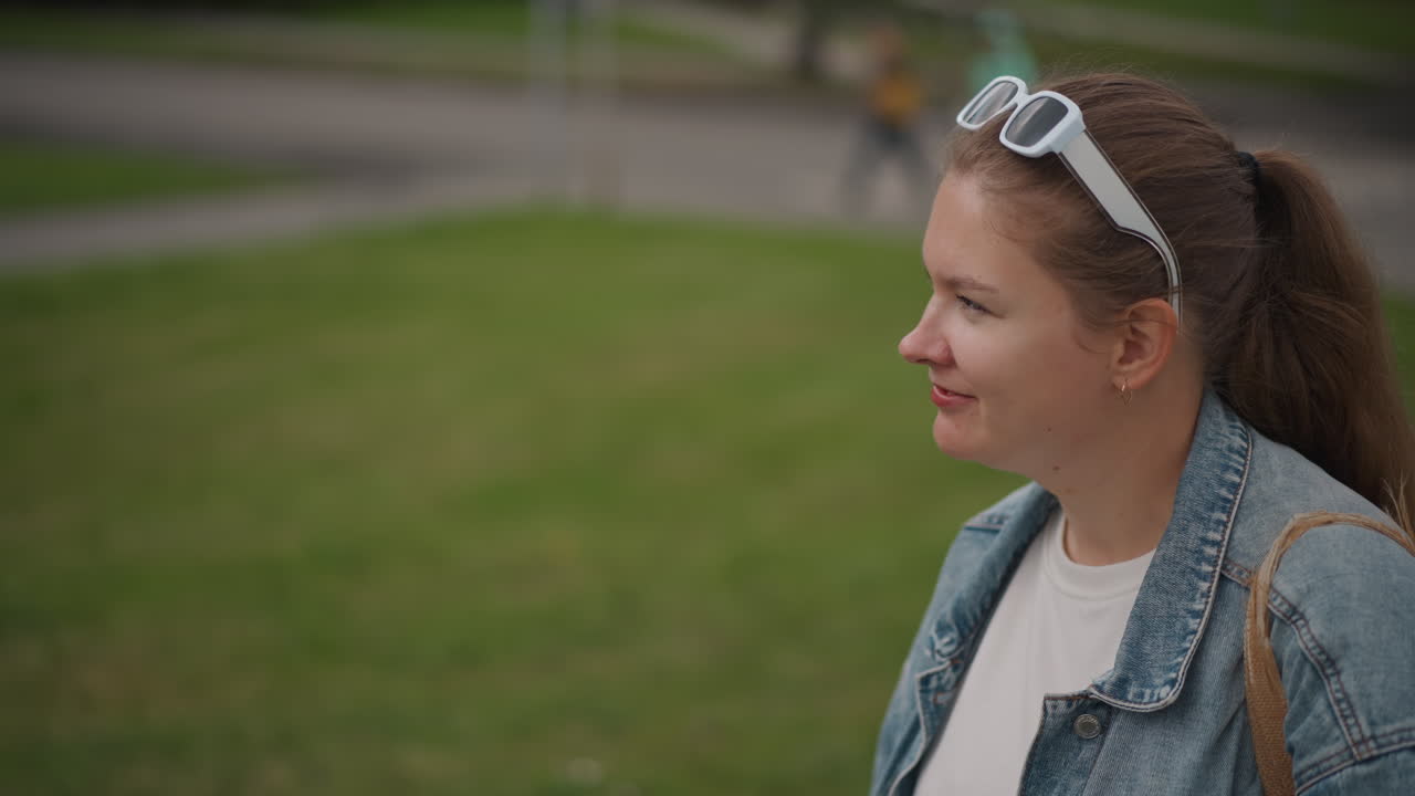 Sideprofil af hvid kvinde i park, denimjakke med solbriller på hovedet, hestehale og blødt smil, ser på sti med afslappet holdning, grøn græsplæne og slørede fodgængere i baggrunden, ægte